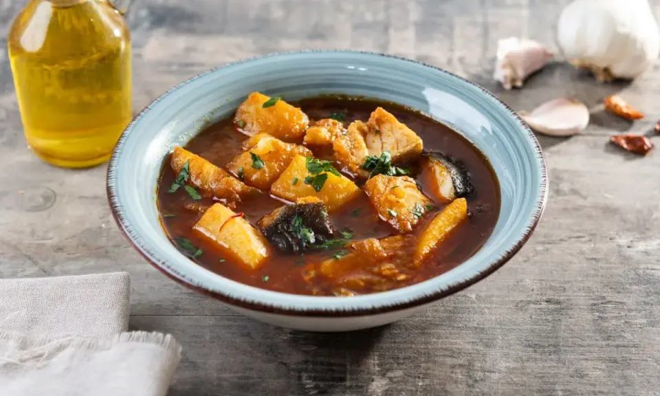 Un tazón de patatas con bacalao en salsa, decorado con perejil. Al lado, una botella de aceite de oliva y ajos.