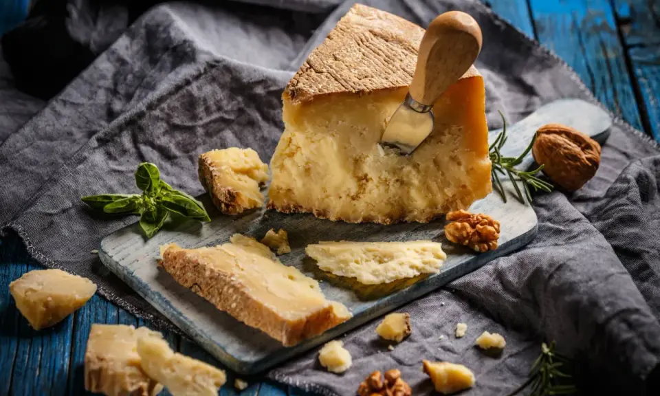 Kawałek parmezanu z nożem, okruchy sera, orzechy i bazylia. Czy parmezan można mrozić? Tak, można go mrozić, choć może stracić nieco na konsystencji.