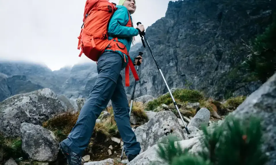 Kobieta z plecakiem wyrusza na wypad w góry, wspierając się kijkami trekkingowymi po kamienistym szlaku.