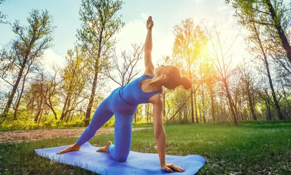 Kvinna utför yoga utomhus i en skogsmiljö. Solen skiner genom träden.