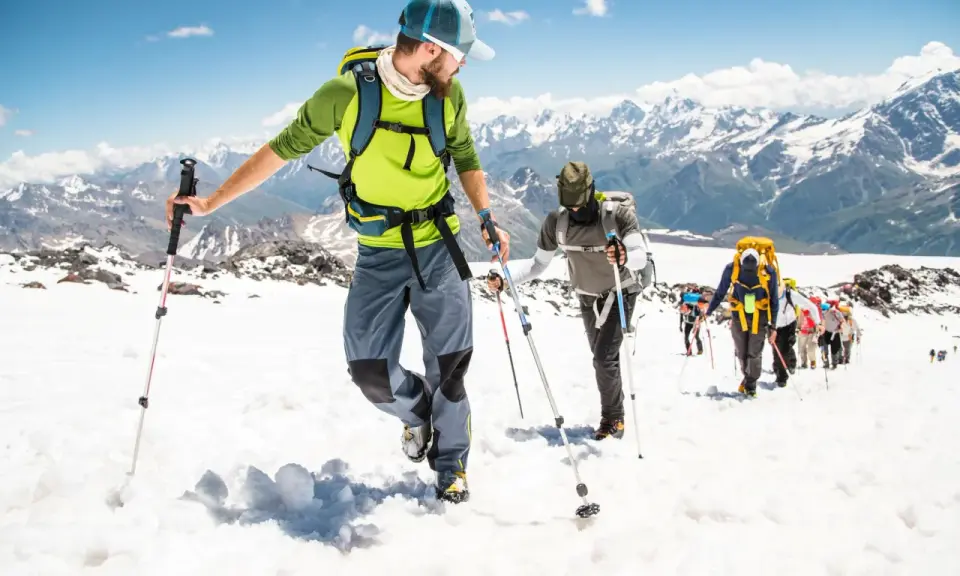 Un gruppo di escursionisti sta camminando sulla neve in montagna, con bastoncini da trekking e zaini, sotto un cielo azzurro.