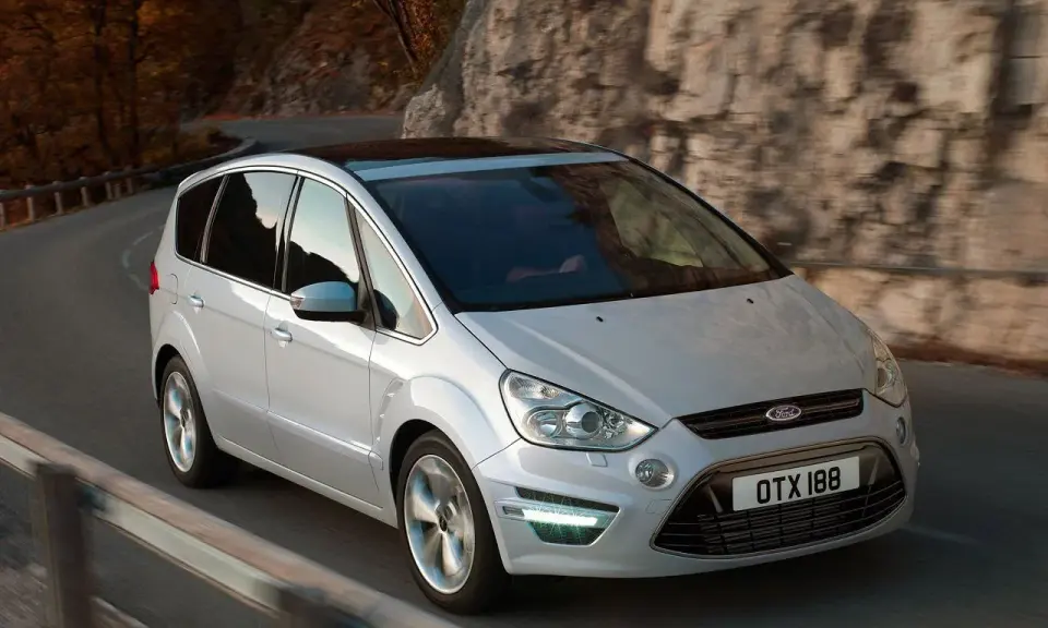 Biały Ford S-Max 2.0 TDCI pokonuje zakręt na malowniczej drodze wśród jesiennych drzew i skał.