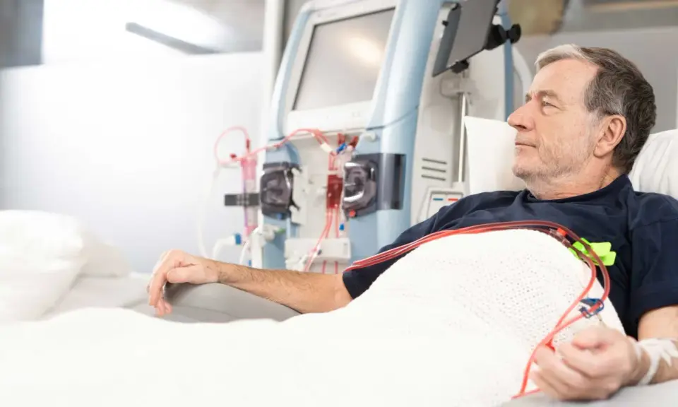 Starszy mężczyzna leży pod kocem, podłączony do aparatury do dializy. Jego ręka spoczywa na podłokietniku, a obok widać maszyny i czerwone wężyki.