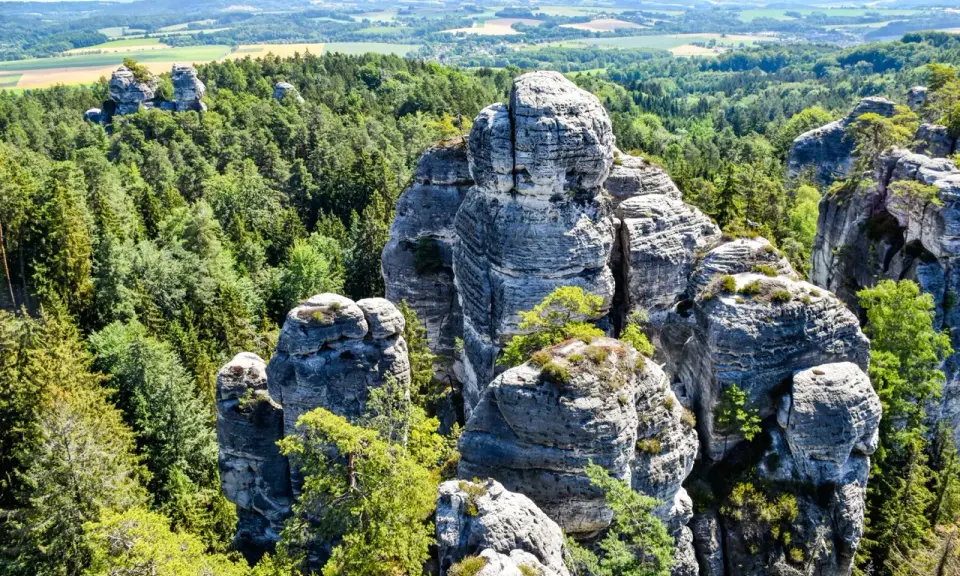Majestatyczna, hruba skala wznosi się pośród gęstego lasu. W oddali widać pagórkowaty krajobraz.