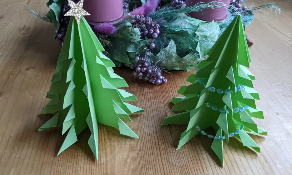 Zwei grüne, flache Origami Tannenbäume stehen auf einem Holztisch. Einer ist mit einer goldenen Sternspitze geschmückt, der andere mit einer Perlenkette. Im Hintergrund ein Adventskranz.