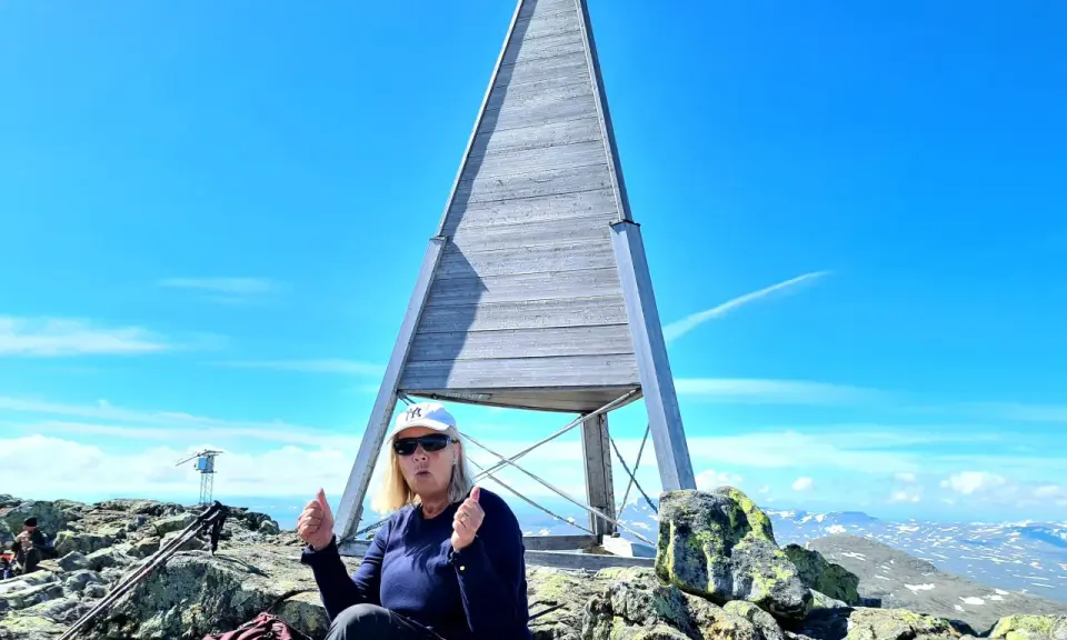 En kvinna sitter på toppen efter att ha lyckats bestiga Helags. Hon ger tummen upp vid den ikoniska träpyramiden.