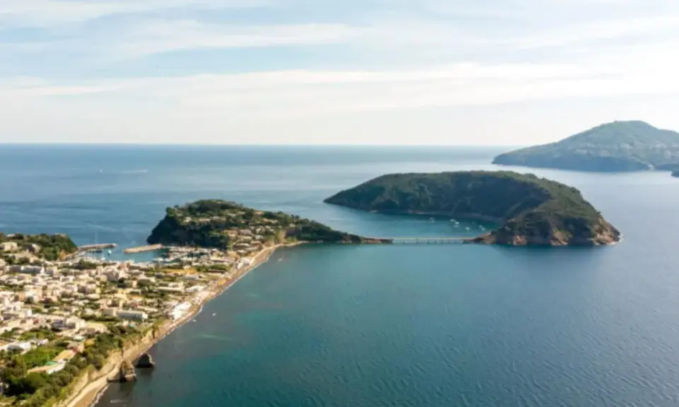 Panoramica di un'isoletta tra Napoli e Ischia, con un borgo costiero, un porto turistico e un ponte che collega a un'altra isola verdeggiante.
