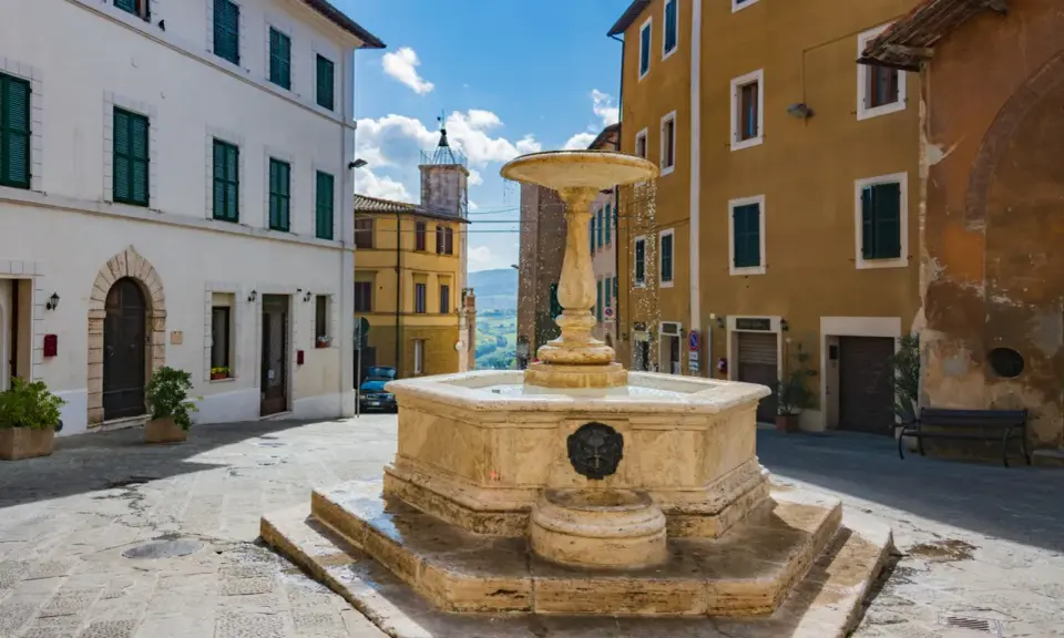 Fontana in pietra nella Valdichiana Senese, circondata da edifici storici con persiane verdi e un cielo azzurro con nuvole bianche.
