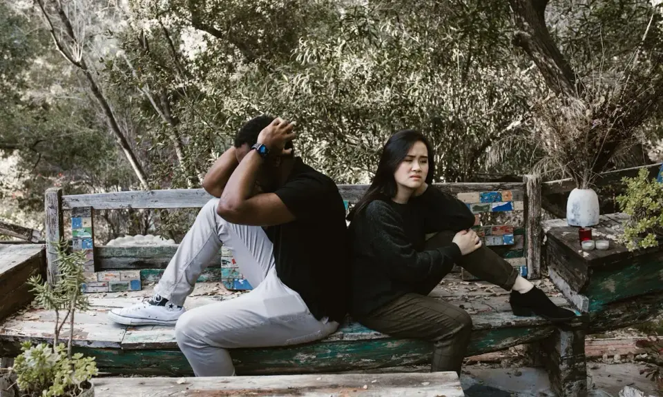 Pareja sentada de espaldas en un banco, con expresiones de angustia. El hombre se agarra la cabeza, ella mira al vacío. Un **vínculo traumático** parece separarlos en este entorno natural.