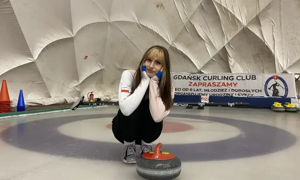 Młoda sportsmenka na lodzie, gotowa do gry w curling. W tle baner "Gdańsk Curling Club Zapraszamy".