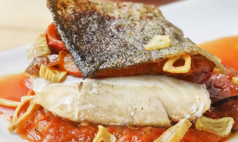 Bacalao a la riojana con piel crujiente, pimientos y ajo frito, servido sobre una cama de salsa de tomate.