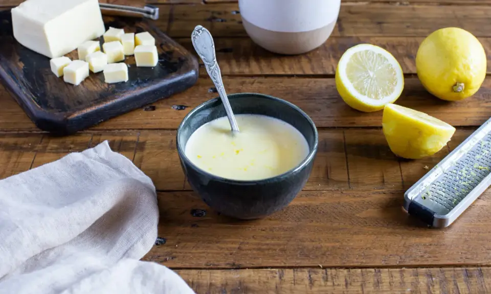 Un bol de sauce beurre citron, avec des citrons, du beurre coupé en dés et une râpe à zester sur une table en bois.