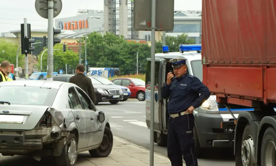 Policjant rozmawia przez radio przy miejscu wypadku. Uszkodzone auto i ciężarówka sugerują, że mogło dojść do zdarzenia związanego z pzu klauzula alkoholowa.