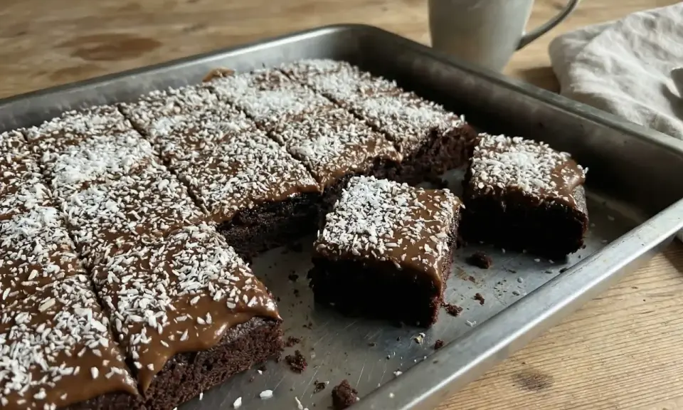 En kladdkaka med glasyr och kokosflingor skuren i bitar i en plåt. En mugg kaffe står bredvid.