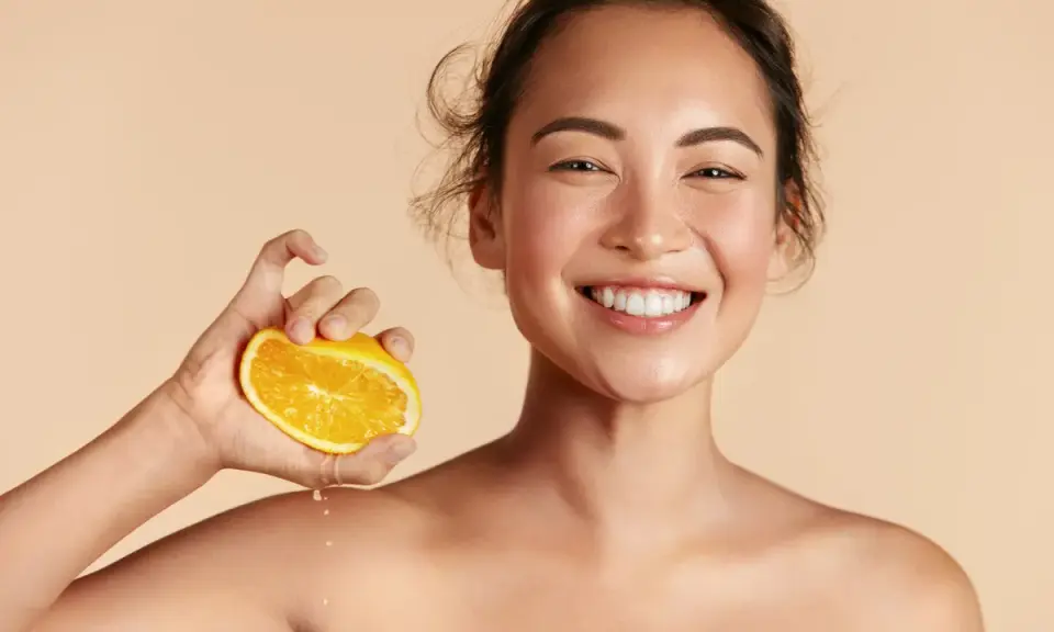 Donna sorridente con fetta d'arancia, suggerisce che il siero alla vitamina C si può usare in estate per una pelle luminosa.