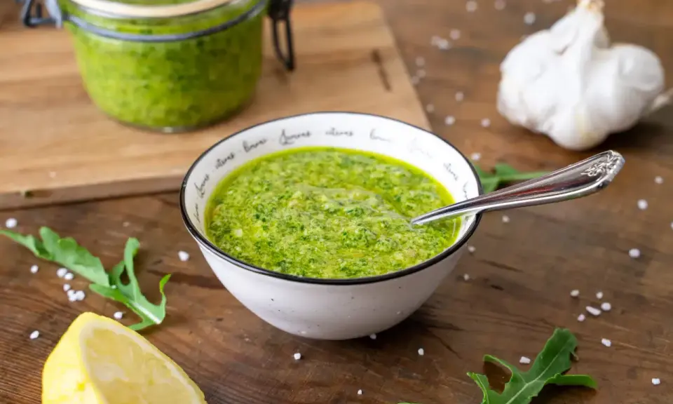 Frisches Rucola Pesto in einer Schüssel, daneben Zitrone, Knoblauch und Rucolablätter.