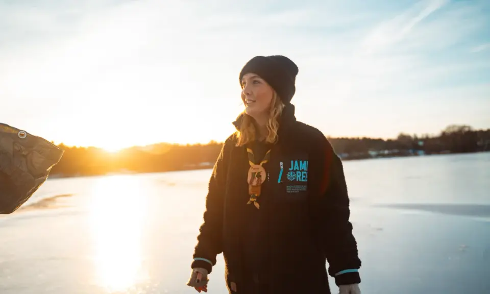 Young woman in a black beanie and fleece jacket standing on a frozen lake at sunset.