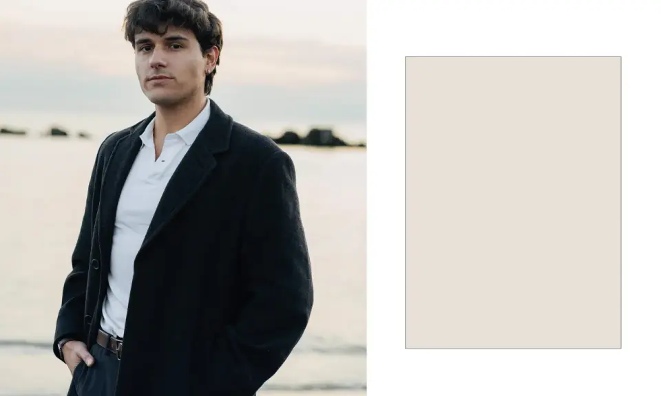 Davide Avolio, con le mani in tasca, indossa un cappotto scuro e una polo bianca, con il mare sullo sfondo.