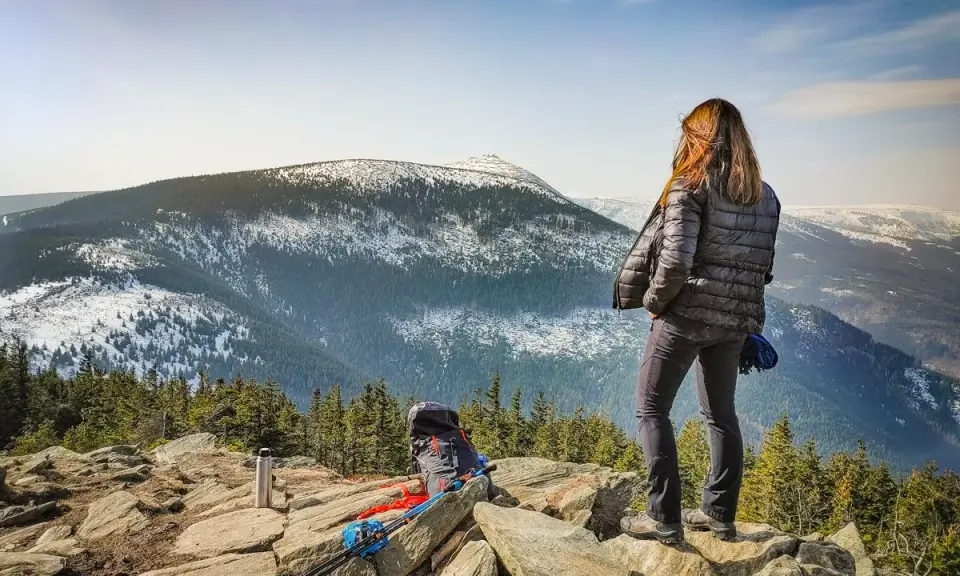 Kobieta na skalnym stole podziwia zimowy krajobraz gór. Obok plecak i kijki trekkingowe.