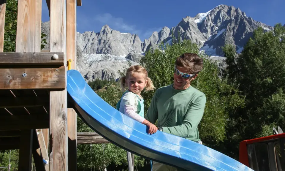 Un padre e una bambina si divertono su uno scivolo in un parco giochi con le maestose montagne della Valle d'Aosta sullo sfondo, un'estate indimenticabile.
