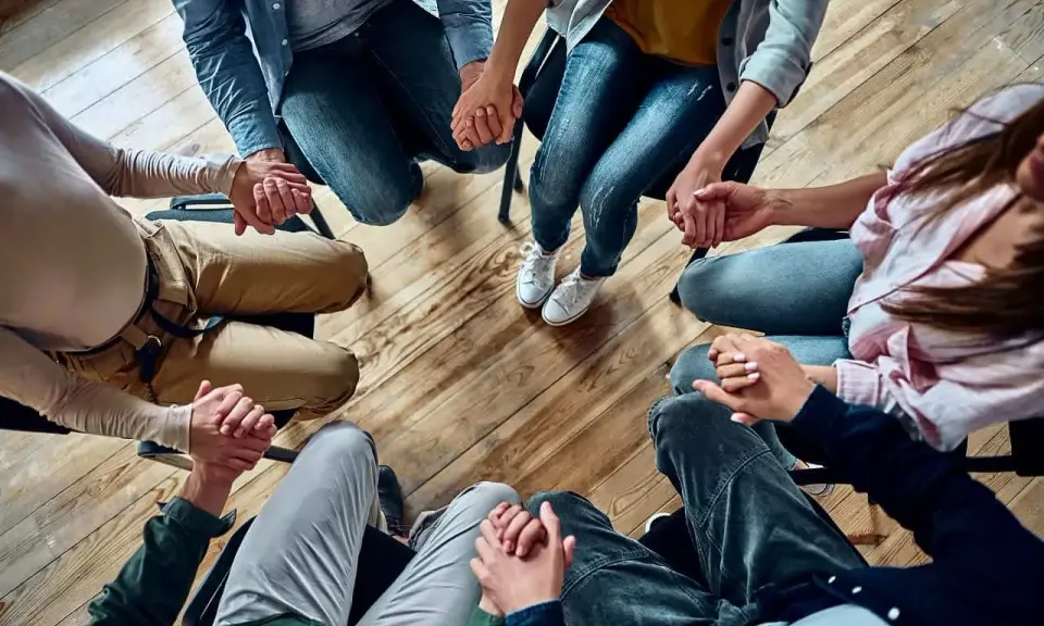 Grupa osób trzyma się za ręce w kręgu, wspierając się nawzajem. Widać tu pracę specjalisty od uzależnień.