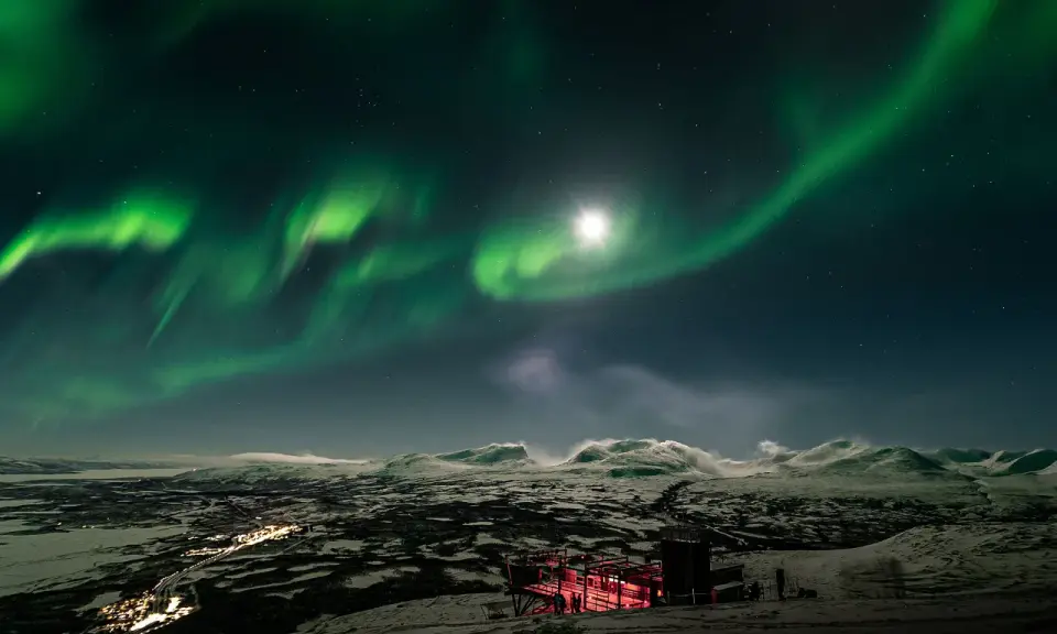 Norrsken över ett snöigt landskap med en upplyst byggnad. Kanske en vy från ett tåg till Abisko.