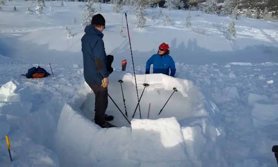 Två personer bygger en bivack i snön. De använder snöblock för att bygga väggarna och stavar för att markera utrymmet.