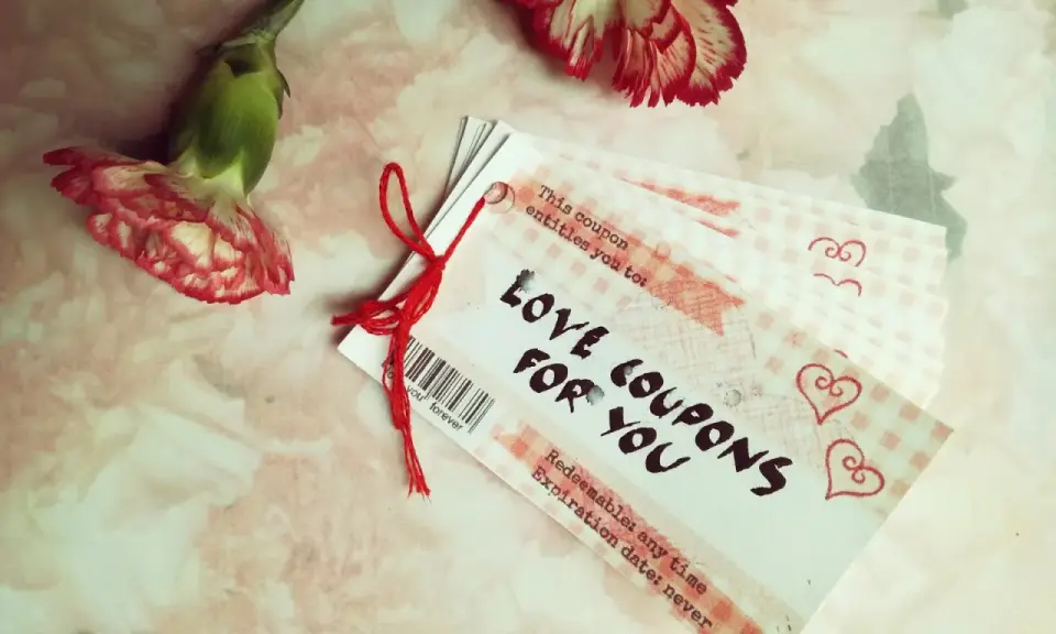 Papierowa rocznica uczczona kuponami miłości. Dwa kwiaty goździka i stosik kuponów "Love Coupons for You" na różowym tle.