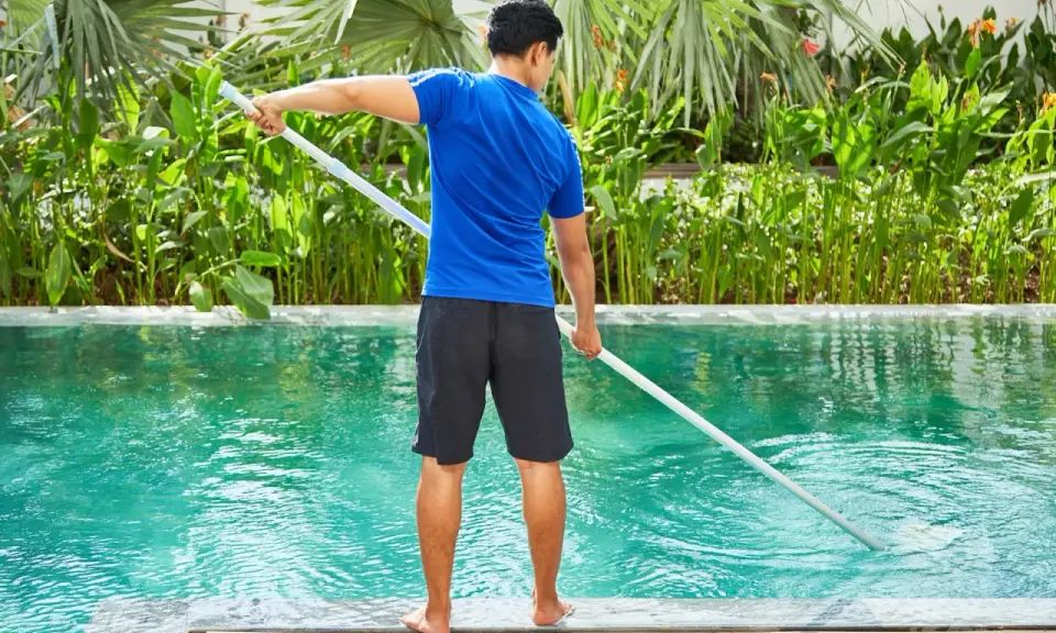 Hombre con camiseta azul y pantalones cortos negros limpia piscina de sal con un limpiafondos.