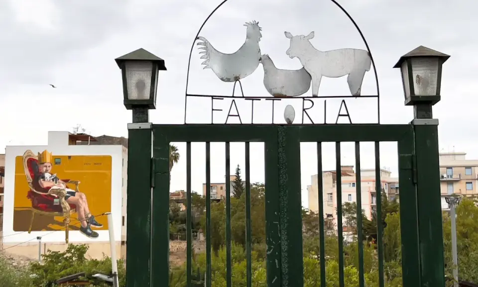Cancello verde con insegna "FATTORIA" e sagome di animali. A sinistra, murale di un uomo su trono. Danisinni, Palermo.