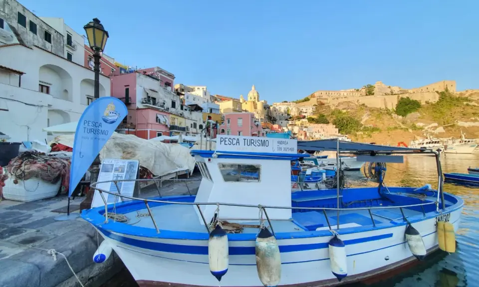 Barca bianca e blu ormeggiata, con bandiera "Pesca Turismo Isola di Procida". Case colorate sullo sfondo.