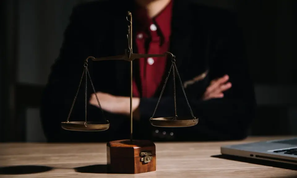 Bilancia della giustizia su un tavolo di legno, con una persona in giacca e camicia rossa sullo sfondo. Le ultime notizie sulla legge Pinto sono in arrivo.