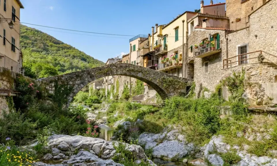Antico ponte in pietra collega i borghi dell'entroterra ligure ponente, con case colorate e vegetazione rigogliosa che scende verso il torrente.