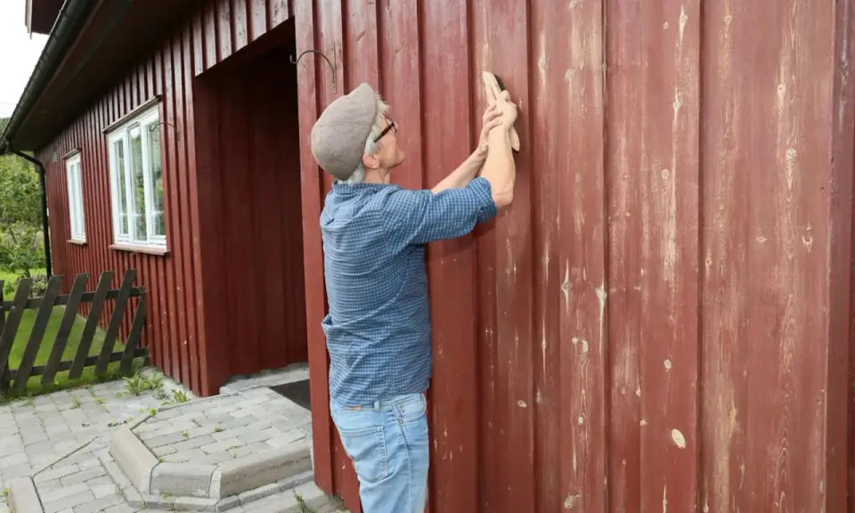 Man slipar träfasaden på ett rött hus. Förberedelser för att måla om.