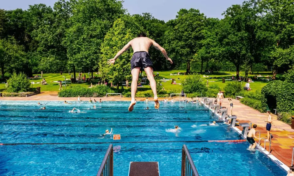 Ein Mann springt in ein Freibad in NRW. Viele Menschen sonnen sich auf der grünen Liegewiese im Hintergrund.
