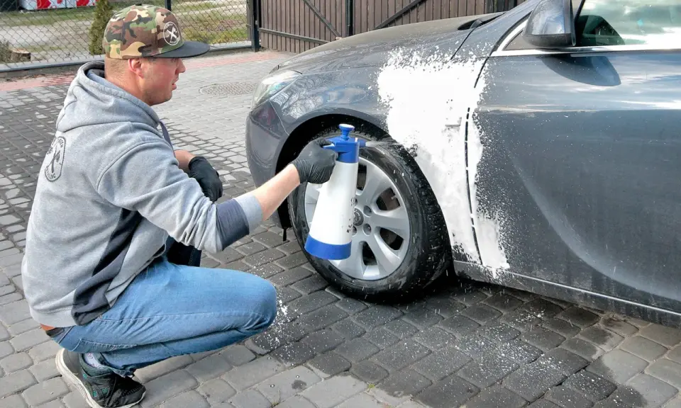 Mężczyzna w czapce z daszkiem i jeansach kuca przy samochodzie, rozpylając pianę na błotnik. Czy można naprawiać auto pod blokiem? Tak, jeśli to drobna naprawa lakieru.