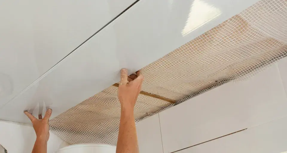 Mains installant des panneaux blancs pour refaire un plafond, avec un treillis visible en dessous.