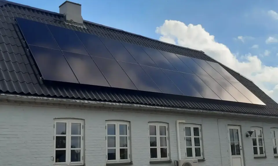 Ett hus med svarta, glänsande nya solceller på taket. Solen reflekteras i panelerna mot en blå himmel med moln.