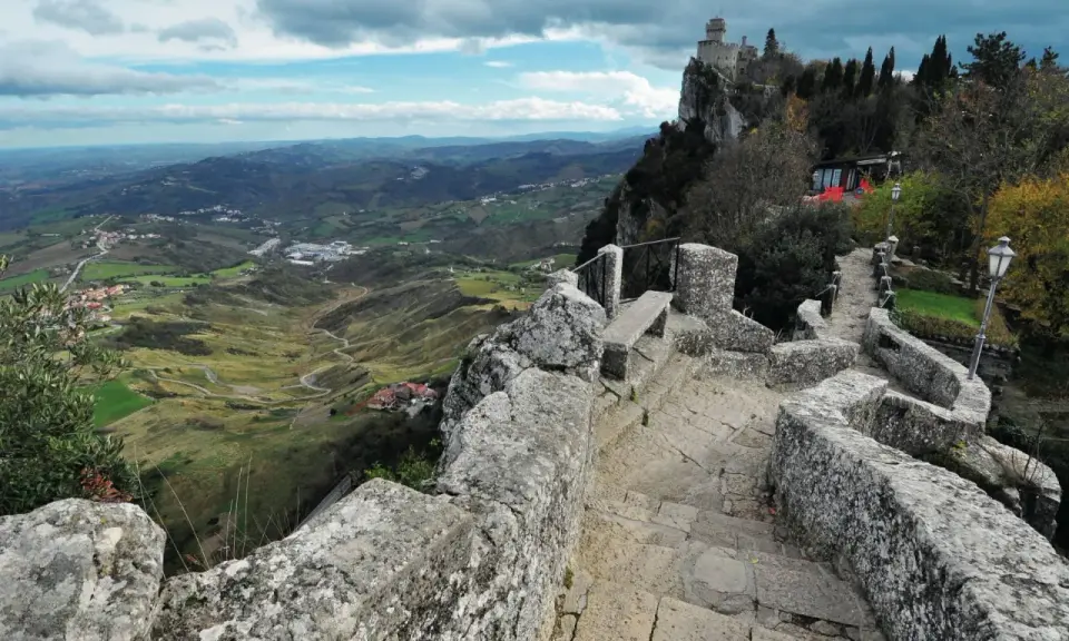 Sentiero in pietra conduce al **San Marino castle**, con vista panoramica sulla valle verdeggiante e colline ondulate sotto un cielo nuvoloso.