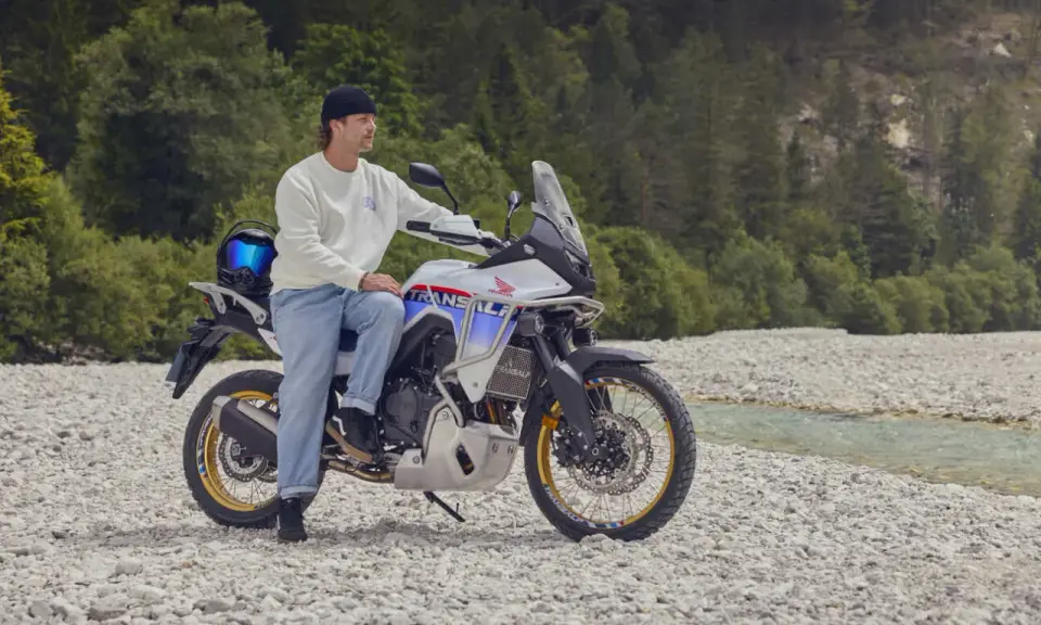 Mężczyzna na motocyklu Honda Transalp, gotowy na przygodę motocyklem po Europie. W tle las i strumień.