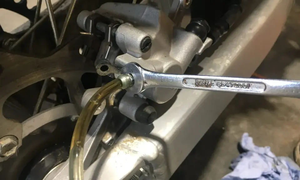 Klucz nasadowy odkręca odpowietrznik zacisku hamulcowego motocykla, podczas gdy przez przezroczysty wężyk płynie płyn. To etap wymiany płynu hamulcowego.