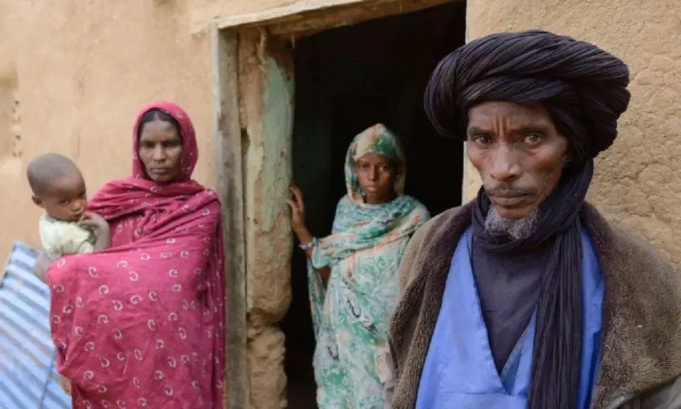 Ein Tuareg-Mann mit schwarzem Turban und ein Kind mit seiner Mutter vor einem Lehmhaus in der Sahara.