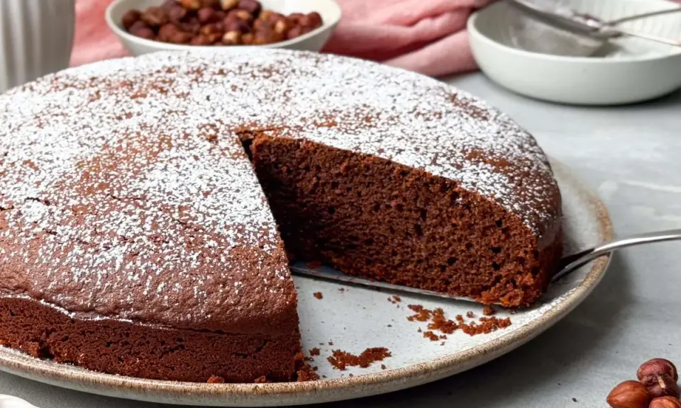 Ein saftiger Nutella Kuchen, bestäubt mit Puderzucker, wartet darauf, angeschnitten zu werden. Daneben eine Schüssel mit Haselnüssen.
