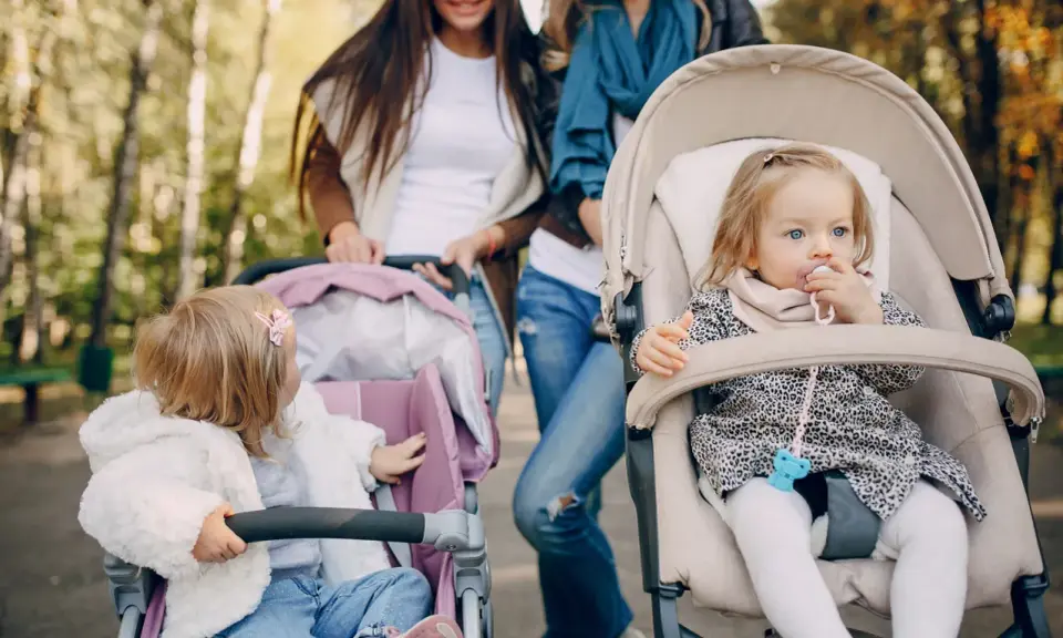 Dwie mamy spacerują z niemowlętami w wózkach. Jedno dziecko ma smoczek, drugie bawi się zabawką. Idealny spacer w parku.