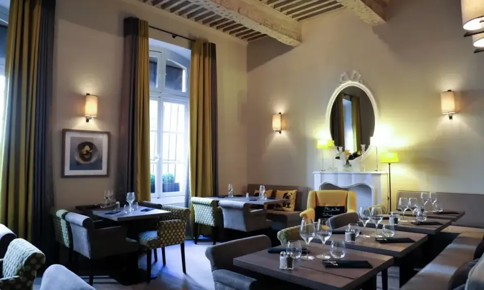 Salle à manger élégante avec tables dressées, prête à servir une spécialité de Provence. Lumière douce, rideaux ocre et gris, miroir orné.
