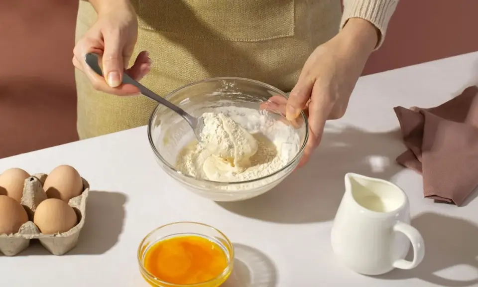 Mani che mescolano farina e burro in una ciotola, con uova, latte e bicarbonato al posto del lievito pronti per la preparazione.