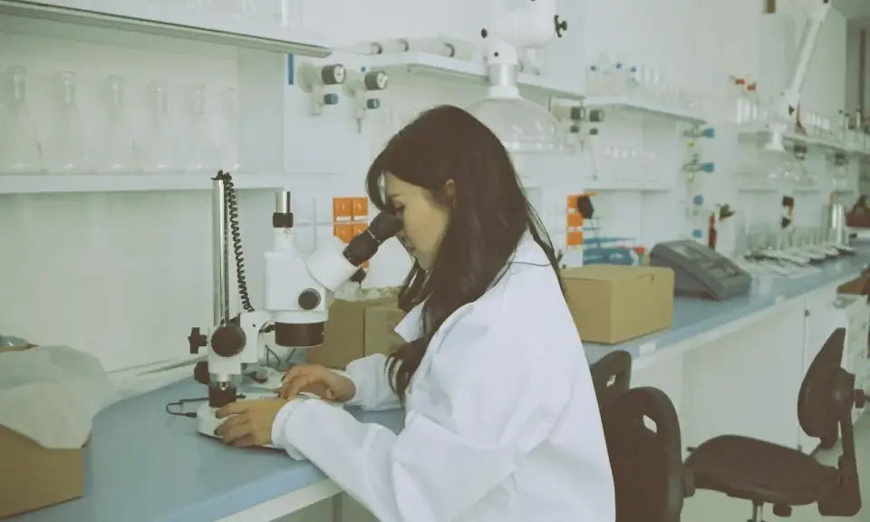 Kobieta w białym fartuchu pracuje przy mikroskopie w laboratorium. Na stole leżą próbki i sprzęt.