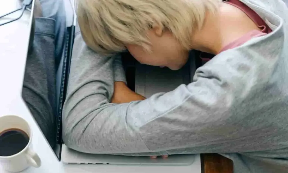 Zmęczona osoba śpi na laptopie. Czy leczenie odwykowe a zwolnienie z pracy to trudny wybór? Kawa obok.