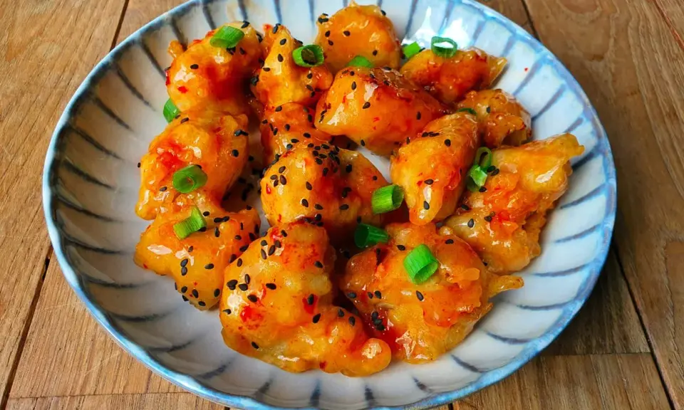 Krispiga bitar av sweet chili kyckling, toppade med sesamfrön och salladslök, serveras på en blårandig tallrik.