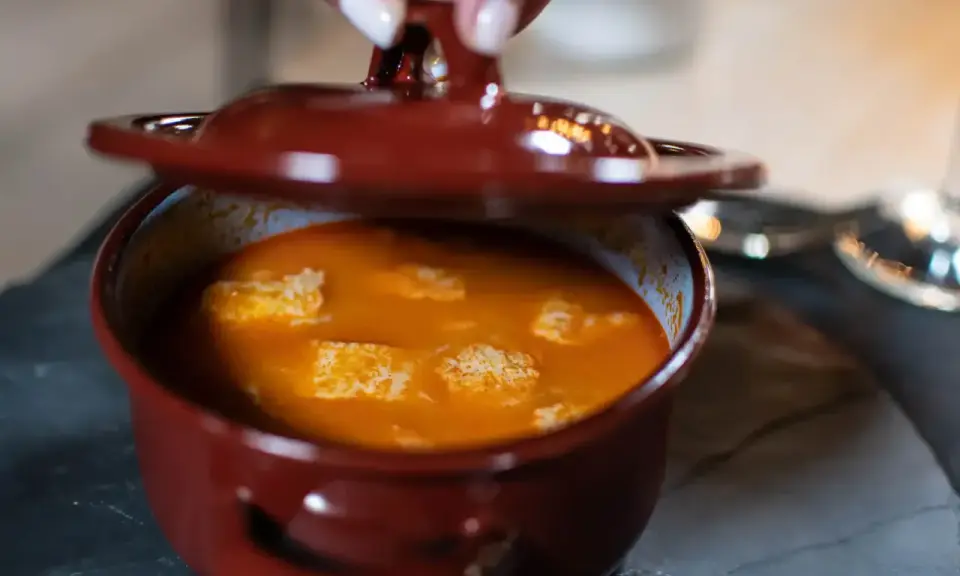 Una mano levanta la tapa de una cazuela de barro con una deliciosa comida típica de Ávila, un guiso naranja con trozos blancos.